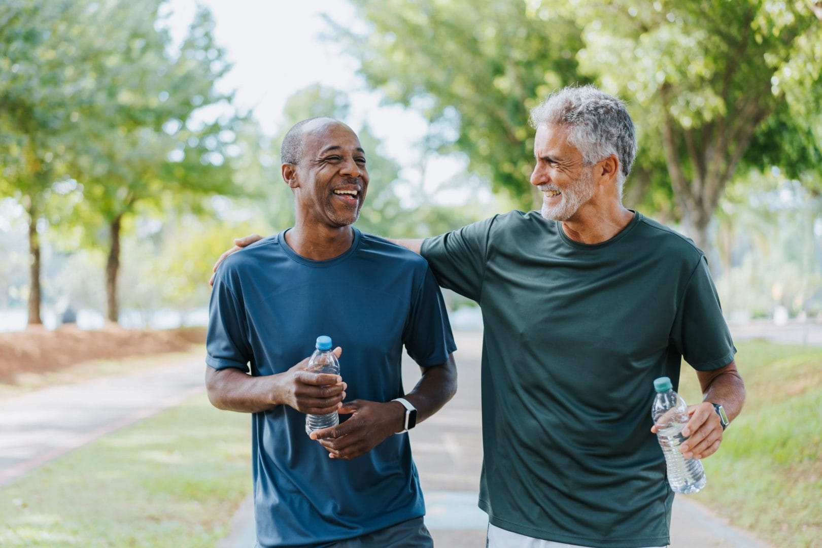 Two active older men walking outdoors, smiling and holding water bottles after a fitness workout.