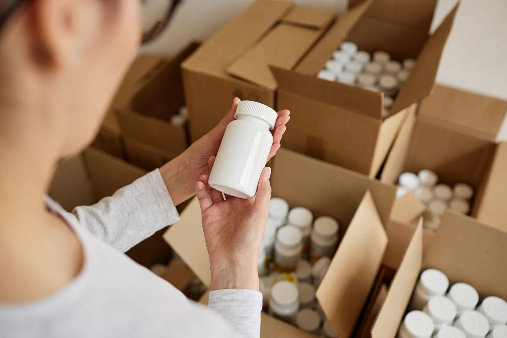Person inspecting a white supplement bottle in a warehouse filled with packaged nutraceutical products.